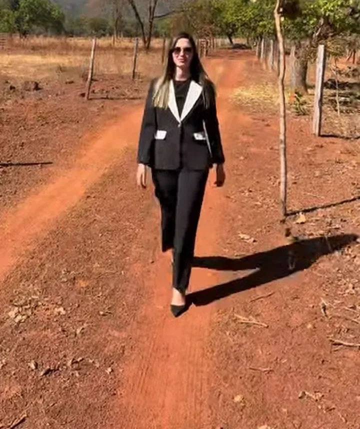 Dra. Laiane, de terno preto com gola branca, caminhando por uma estrada de terra vermelha entre cercas de arame.