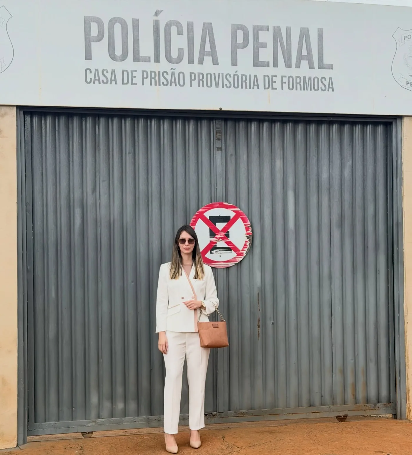 Dra. Laiane Lourenço, de terninho branco e bolsa caramelo, diante do portão de aço da Casa de Prisão Provisória de Formosa.