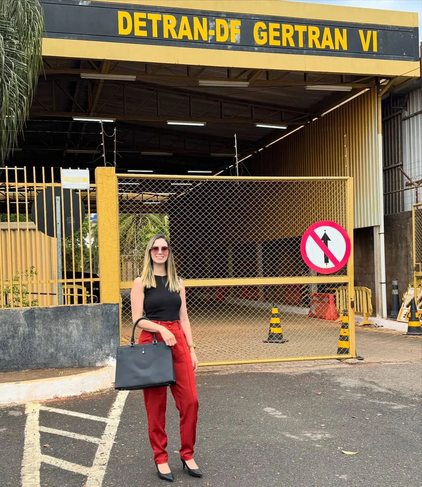 Dra. Laiane Lourenço, de blusa preta, calça vermelha e bolsa preta, diante do portão amarelo do Detran-DF, Gertran VI.