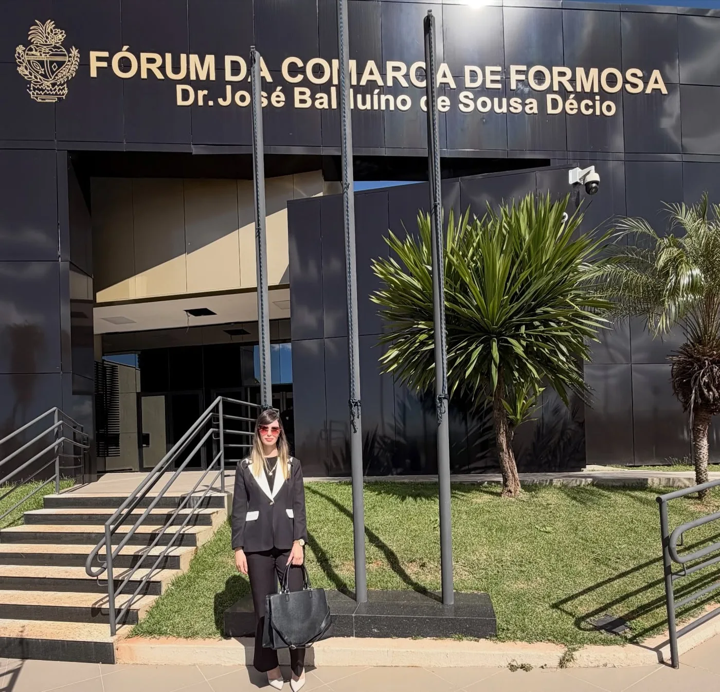 Dra. Laiane Lourenço, de blazer preto e branco e bolsa preta, de pé diante da escadaria do Fórum da Comarca de Formosa.
