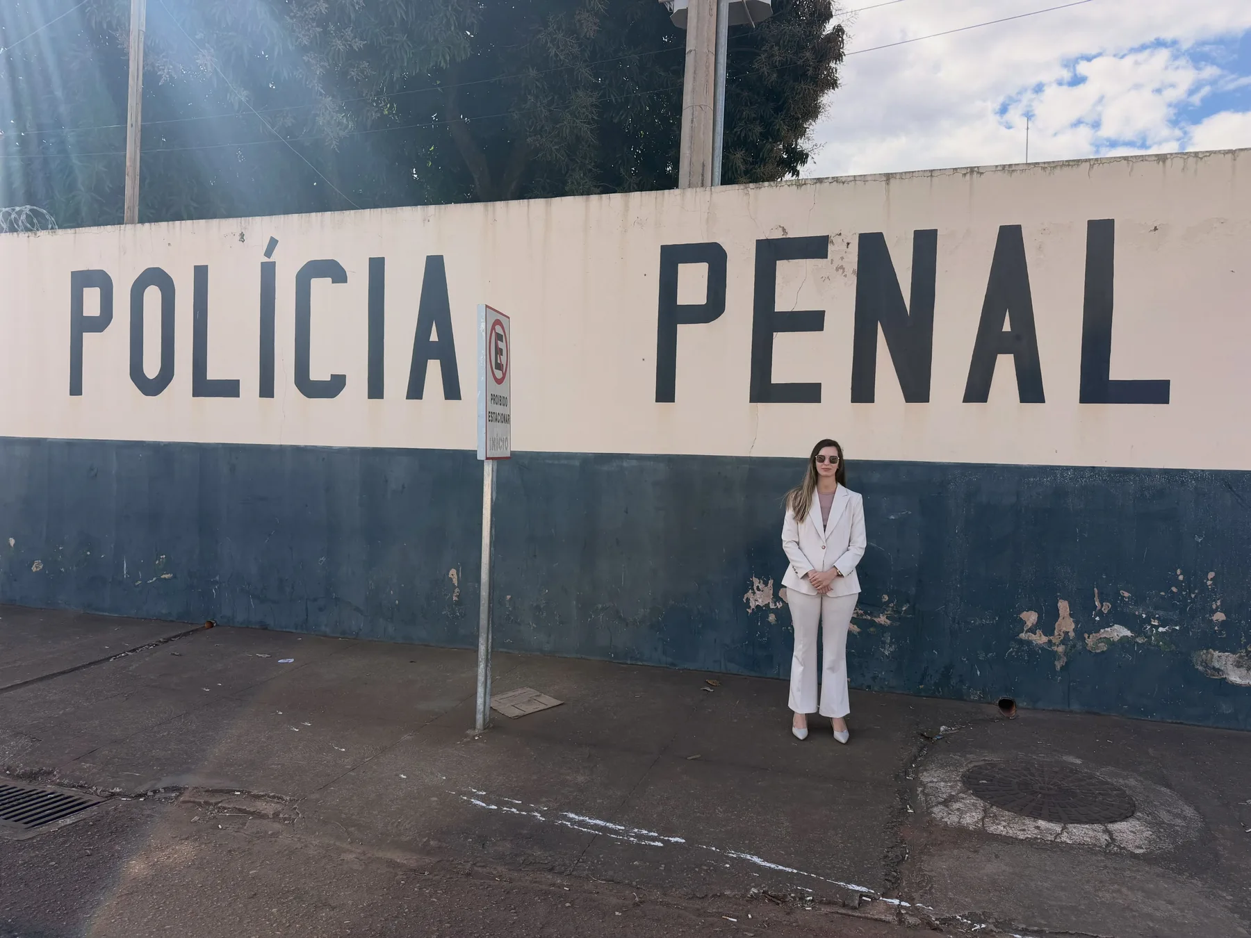 Muro azul e branco com a inscrição POLÍCIA PENAL; a Dra. Laiane, de terninho branco, de pé junto ao muro.