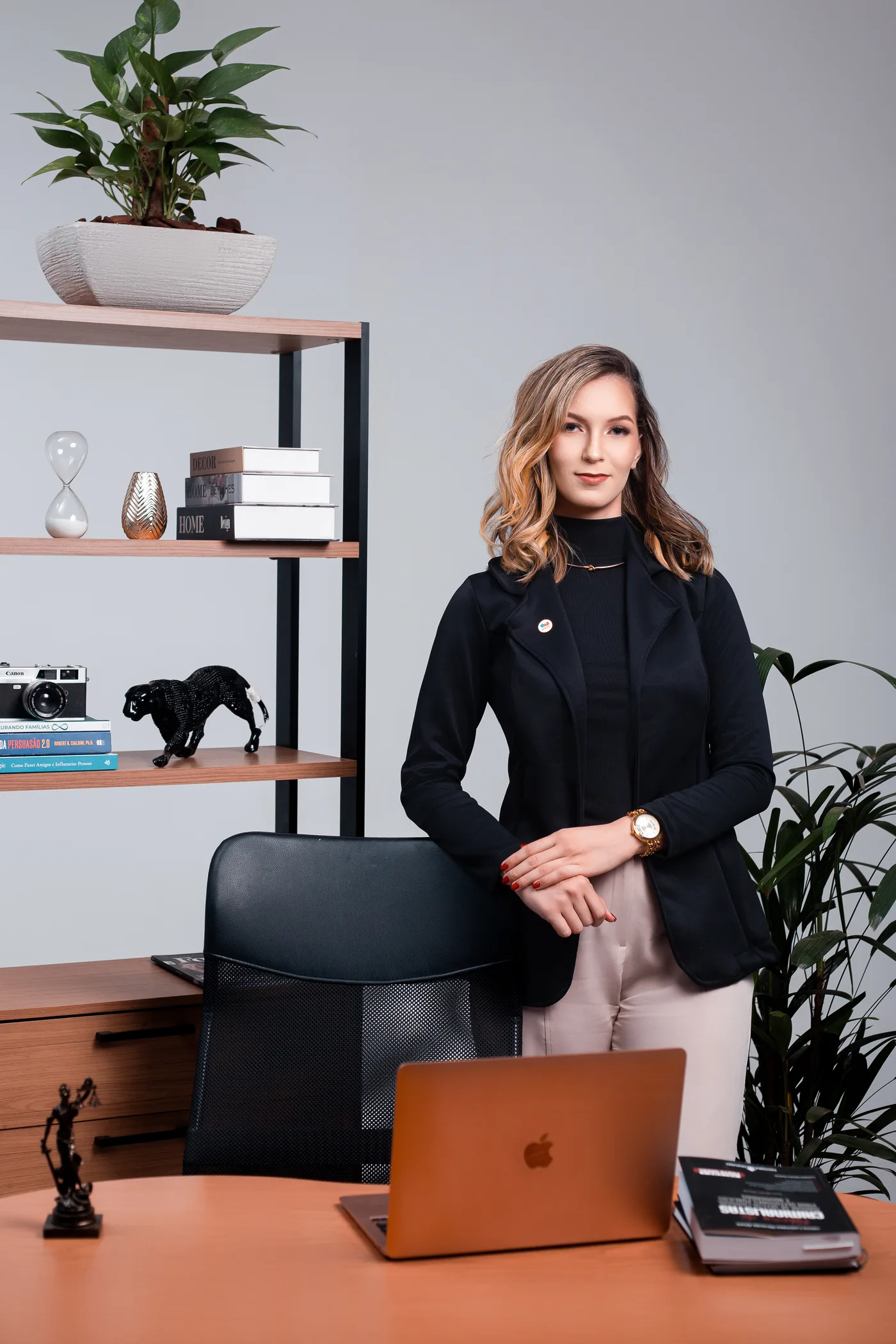 Dra. Laiane Lourenço de pé atrás da mesa do escritório, de blazer preto, mãos cruzadas, com uma estante ao fundo.